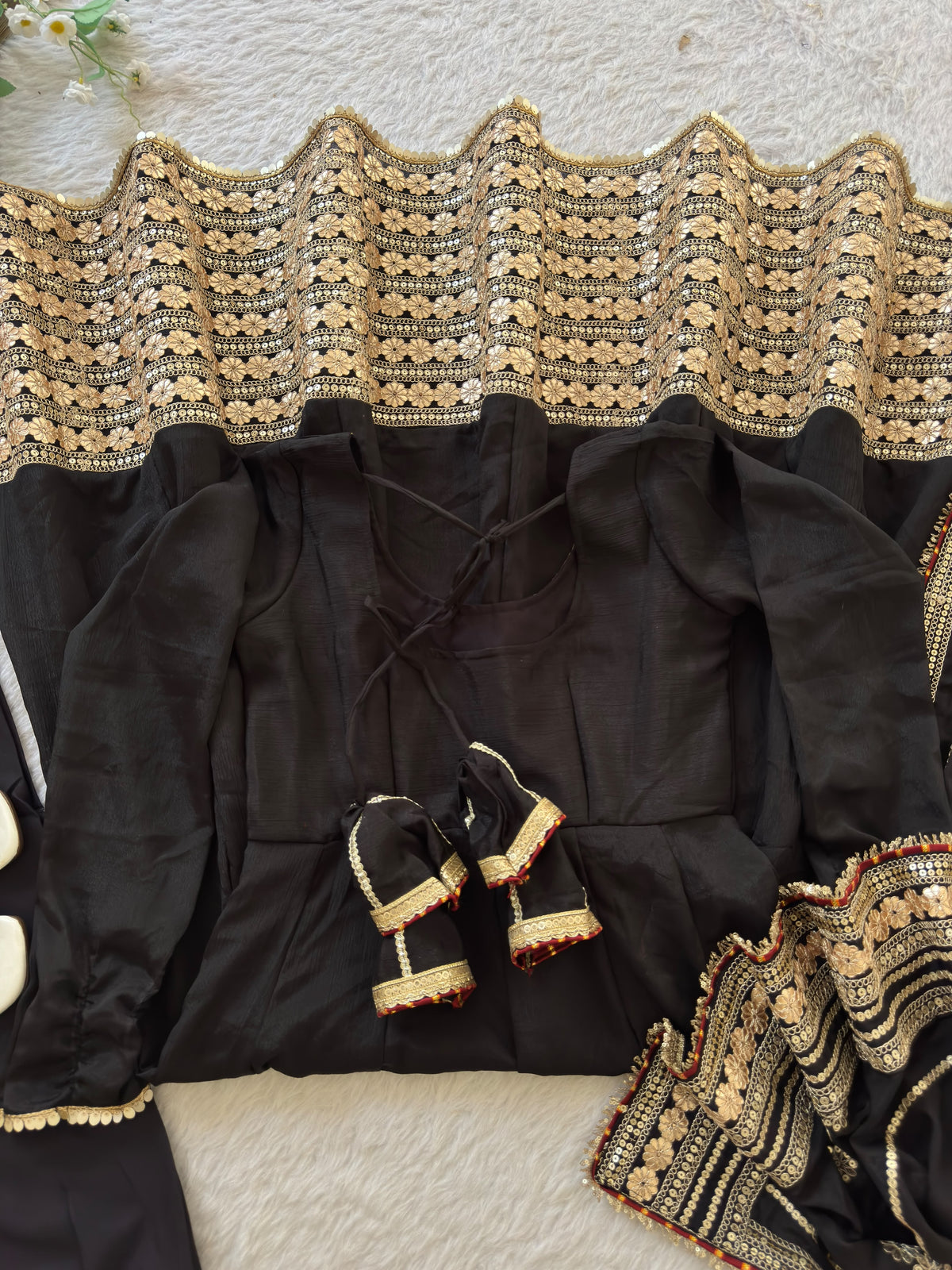 Midnight Black Chinon Silk Anarkali Gown Set - flat lay product close-up showing complete set with gown back tie-back criss-cross lace-up detail, full sleeves with gold lace cuff and gold zari trim, distinctive scalloped gold zari edge on wide dense gold sequence floral daisy hem border, crepe pant and matching dupatta