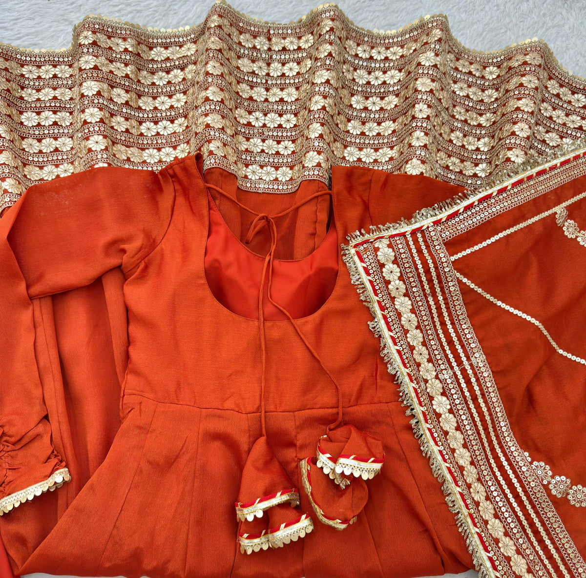 Burnt Orange Rust Chinon Silk Anarkali Gown Set - flat lay close-up showing gown top with round scoop neck, back keyhole tie detail, full sleeves with gold coin lace cuff trim, spectacular scalloped gold sequence floral daisy hem border, and matching multi-stripe dupatta with cream gold floral embroidery stripe bands and scalloped gold border