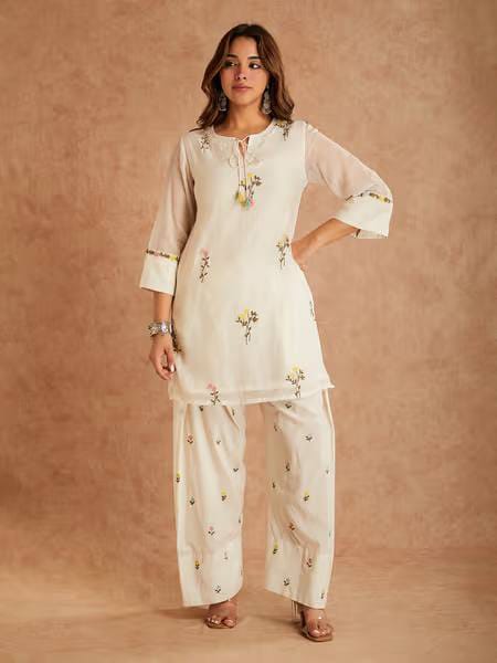 Off-White Cotton Silk Top Farshi Salwar Set - home flat lay overhead view on white polka dot fabric showing complete set with top displaying scattered multicolour wildflower embroidery, tassel tie neckline, sheer bell sleeves, farshi salwar with elasticated waist and matching wildflower butti embroidery, and organza dupatta with pink polka dot digital print and pearl lace border alongside potted plant