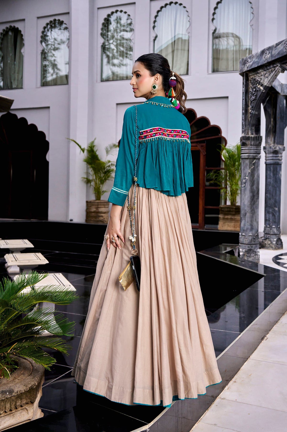 Teal Beige Pure Cotton Lehenga Set - indoor heritage terrace close-up seated model view showing teal crop jacket upper body with open-front lapel collar, gold bead ghungroo fringe drop trim on lapel and front placket, white ikat stripe patch pockets, sheer panel detail, beige blouse inner, and beginning of 8-metre flare beige lehenga