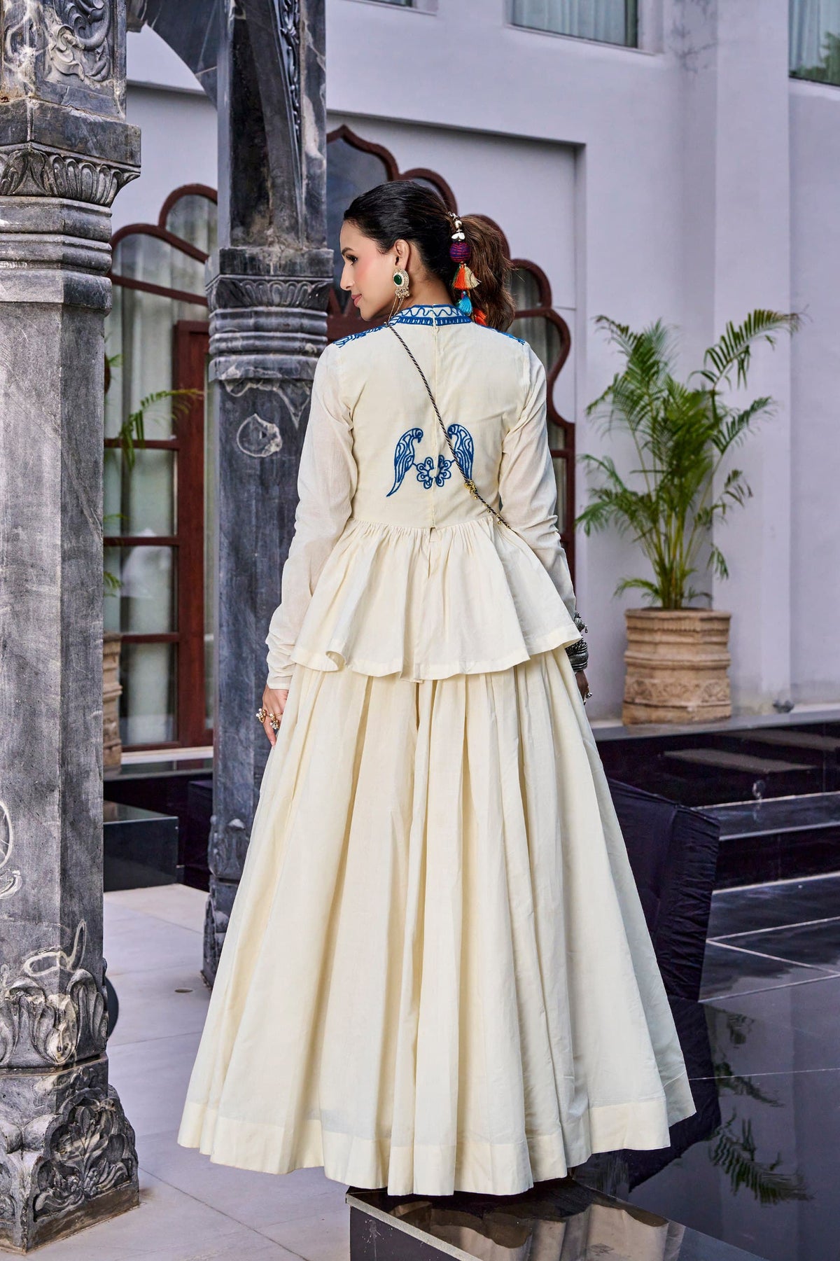 Off-White Royal Blue Kashida Gamthi Lehenga Set - heritage haveli exterior full length back standing model view showing back of off-white peplum top with blue peacock bird pair Kashida motif centrally placed above double-tiered gathered peplum flair, sheer full organza sleeves, and 8-metre full flare off-white cream lehenga with cream hem border