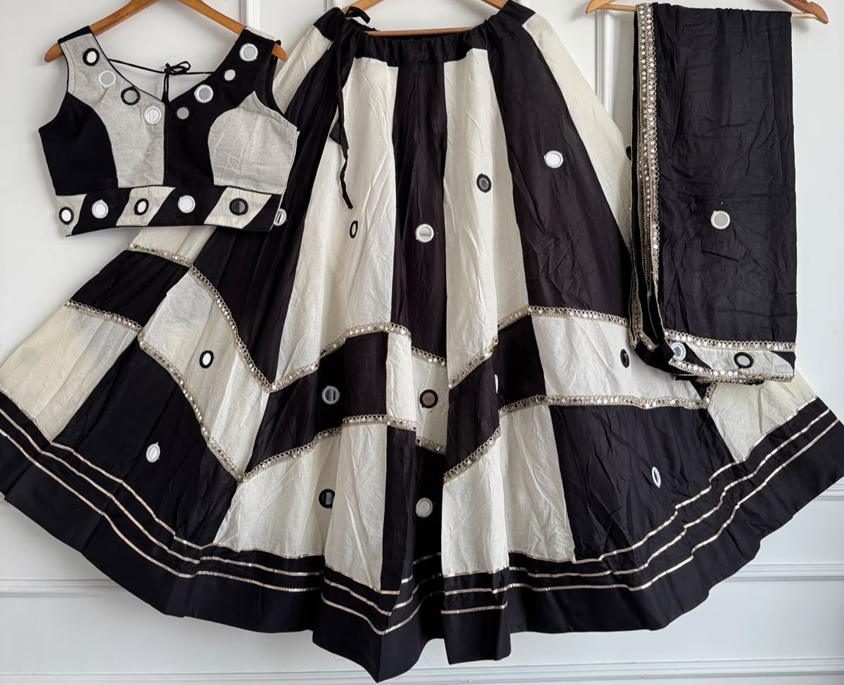 Black White Pali Cotton Chaniya Choli Set flat lay hanger showing complete 3-piece: colour-block blouse with real mirror work and V-neck, chevron block lehenga with diagonal black off-white panels, real circular mirror butti, gold sequence lace tier borders, triple gold zari hem stripe, and black rayon dupatta with mirror butti