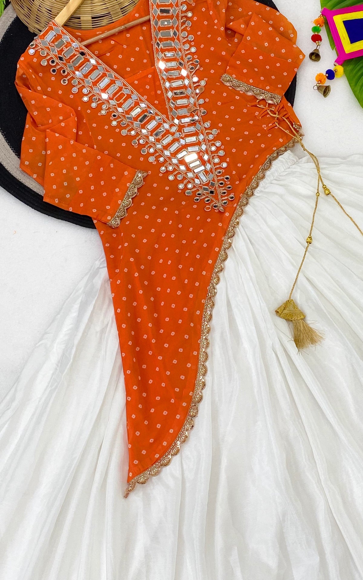 Orange Fox Georgette Asymmetric Kurti White Lehenga Set - overhead flat lay close-up detail showing silver mirror grid yoke panel with large rectangular oval mirrors and scattered round silver mirrors on bandhani butti georgette, scallop gold lace trim on asymmetric hem edge, gold braided cord mustard tassel latkan, and white lehenga fabric
