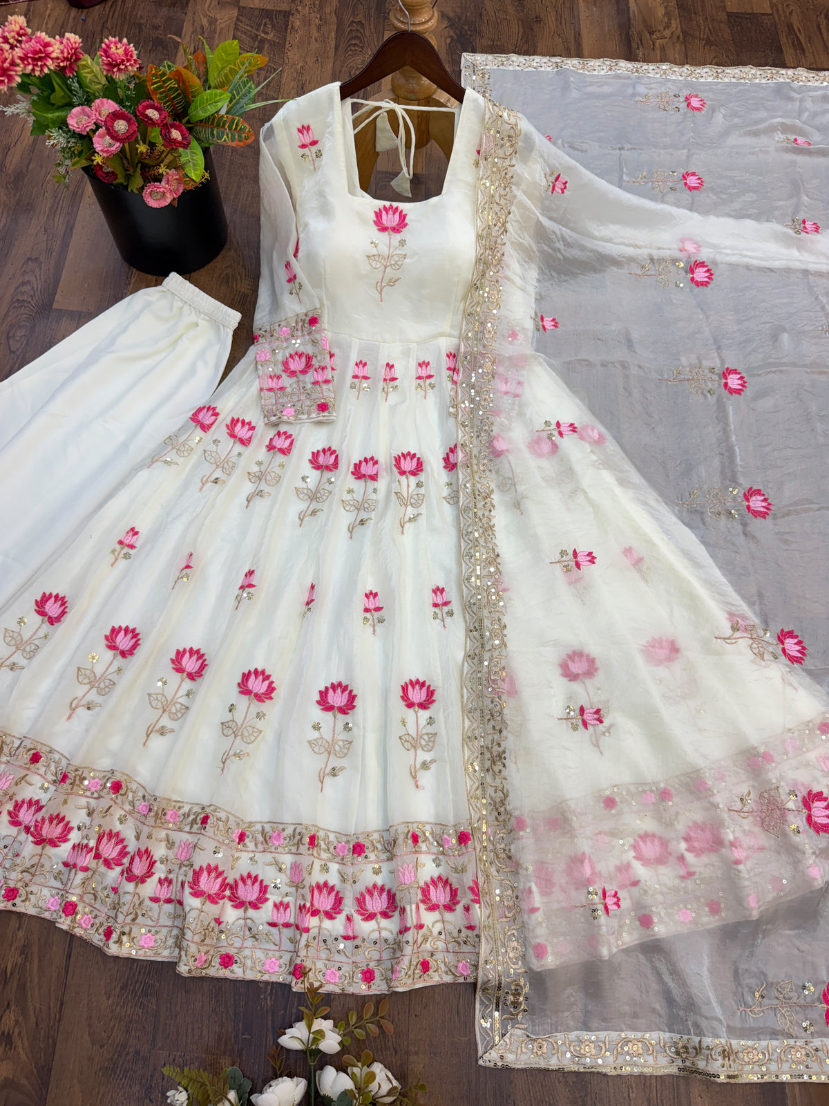 Off-White Fendi Silk Anarkali Gown Set - flat lay hanger dark wood background close-up showing square neckline with gold zari trim, back tie-up criss-cross lace detail, full sheer net sleeves with scattered lotus embroidery, all-over hot pink lotus flower embroidery body with gold zari stems and leaves, and gold sequence lace cuff band on sleeve