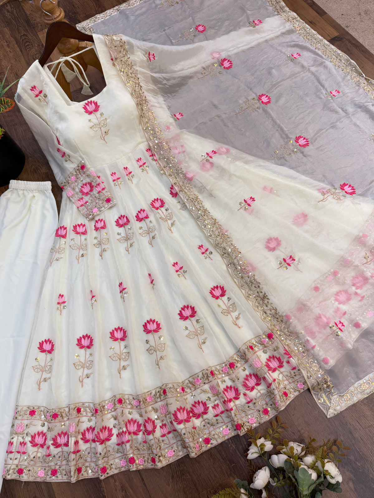 Off-White Fendi Silk Anarkali Gown Set - flat lay dark wood background full complete set overhead view showing gown with all-over lotus embroidery, spectacular wide multi-row lotus floral hem border with graduating hot pink and blush lotus rows and gold sequence bands, white micro cotton straight pant and net dupatta with scattered pink lotus butti and wide gold sequence scrollwork border