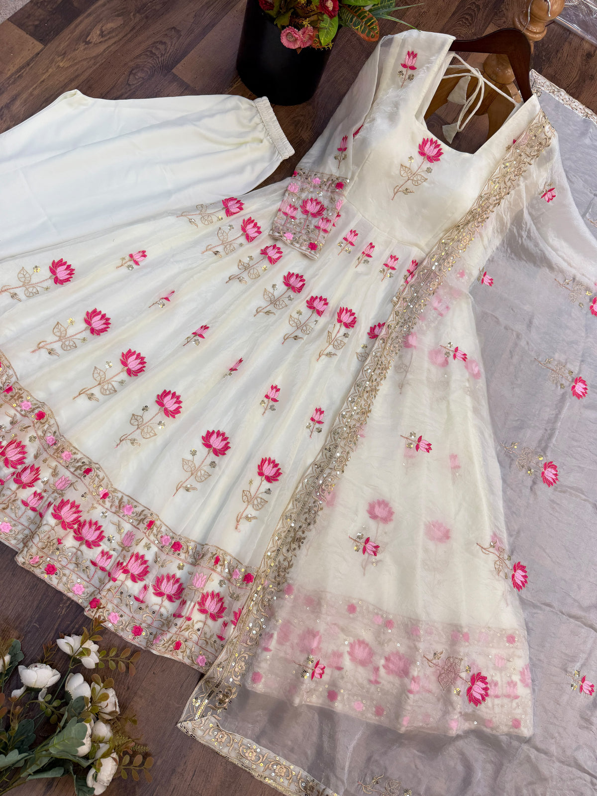 Off-White Fendi Silk Anarkali Gown Set - flat lay dark wood background upper-body close-up showing top section of gown with back tie-up criss-cross halter lace detail, square neckline, all-over scattered pink lotus flower embroidery on ivory fendi silk body, net sleeves with lotus embroidery, and matching net dupatta with scattered pink lotus butti and gold sequence border