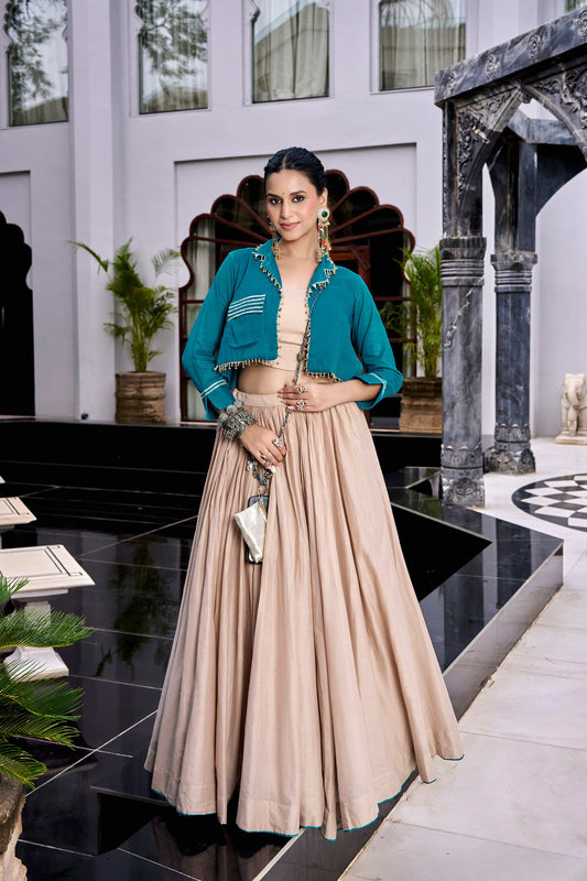 Teal Beige Pure Cotton Lehenga Set - luxury heritage haveli exterior full length standing model front view showing complete set with teal crop jacket with gold bead ghungroo fringe drop trim on lapel and hem, white ikat stripe patch pockets and sleeve cuffs, beige 8-metre flare lehenga with teal hem border, beige blouse inner, and gold chain structured clutch purse